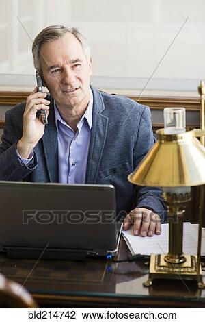 Stock Image - Caucasian businessman talking on cell phone at desk. Fotosearch