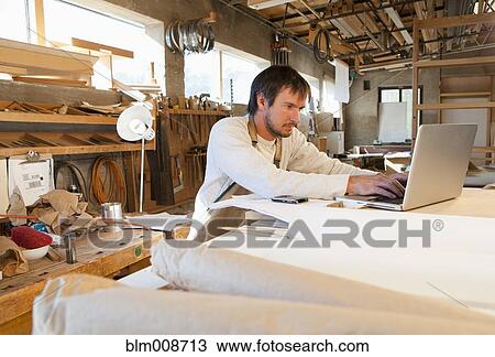 Stock Image - Caucasian carpenter using workshop on laptop. Fotosearch