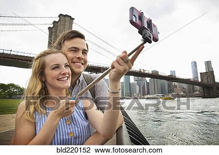 Stock Image - Caucasian couple taking selfie with cell phone under Brooklyn Bridge, Brooklyn, New York, United States. Fotosearch
