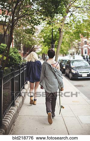 Stock Photograph - Caucasian couple walking on city sidewalk with skateboard. Fotosearch