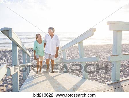 Stock Image - Caucasian couple walking on wooden boardwalk on beach. Fotosearch
