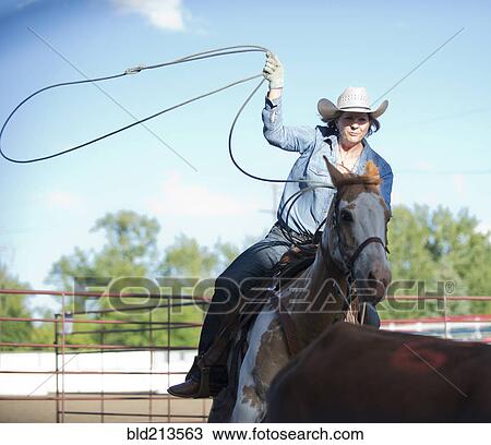 Stock Image - Caucasian cowgirl on horse throwing lasso on ranch. Fotosearch