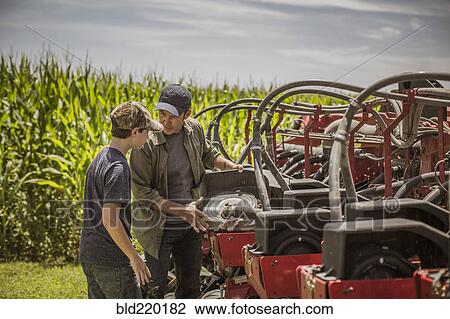 Stock Image - Caucasian farmer explaining machinery to son on farm. Fotosearch
