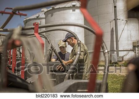 Stock Image - Caucasian farmer explaining machinery to son on farm. Fotosearch