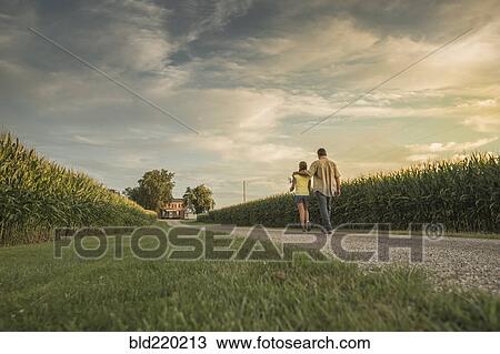 Stock Image - Caucasian father and daughter walking on dirt path by corn field. Fotosearch