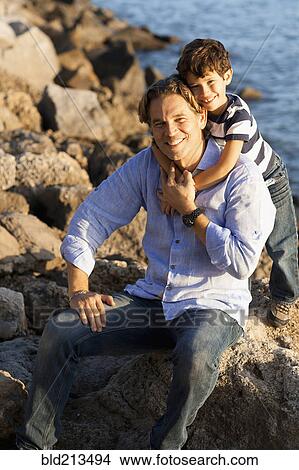 Picture - Caucasian father and son hugging on rocky beach. Fotosearch