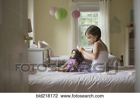 Stock Image - Caucasian girl brushing hair of doll on bed. Fotosearch