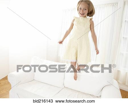 Stock Image - Caucasian girl jumping on sofa in living room. Fotosearch