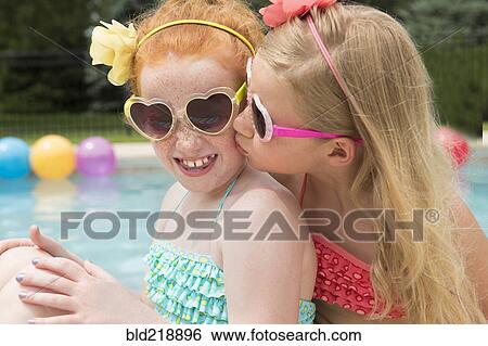 Stock Photograph - Caucasian girl kissing cheek of friend near swimming pool. Fotosearch