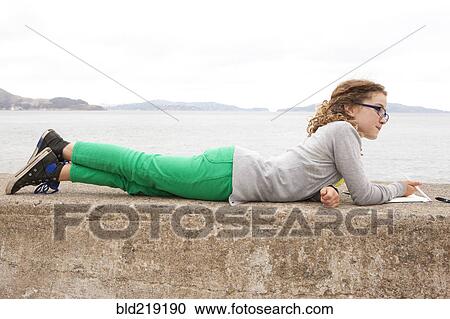 Stock Image - Caucasian girl laying on concrete wall over ocean. Fotosearch