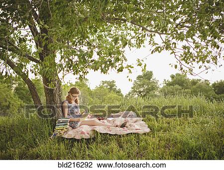 Stock Image - Caucasian girl reading book under tree in field. Fotosearch