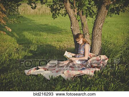 Stock Image - Caucasian girl reading book under tree in field. Fotosearch
