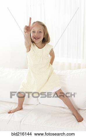 Picture - Caucasian girl standing on sofa gesturing peace sign. Fotosearch