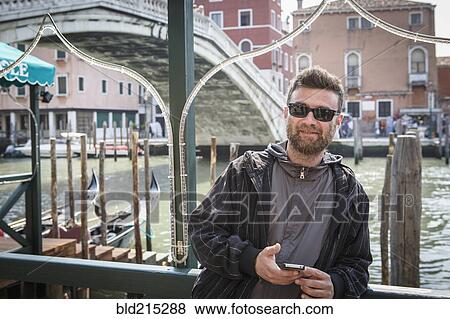 Stock Photo - Caucasian man holding cell phone on dock, Venice, Veneto, Italy. Fotosearch