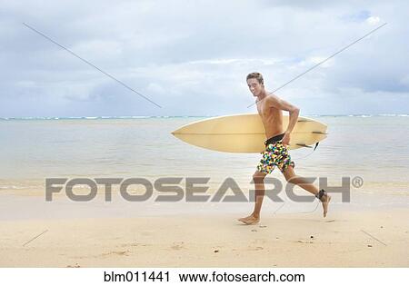Stock Image - Caucasian man running on beach carrying surfboard. Fotosearch