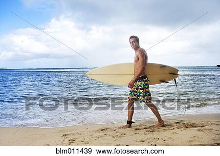 Stock Photo - Caucasian man walking on beach carrying surfboard. Fotosearch