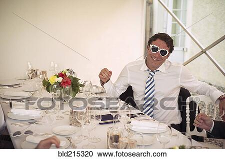 Stock Image - Caucasian man wearing heart-shaped glasses at table. Fotosearch