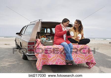Stock Image - Caucasian mother and daughter eating picnic in car trunk on beach. Fotosearch