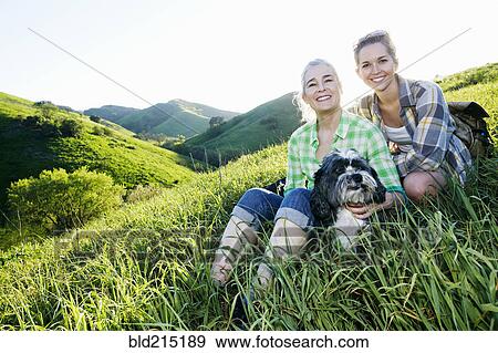 Stock Photo - Caucasian mother and daughter petting dog on grassy hillside. Fotosearch