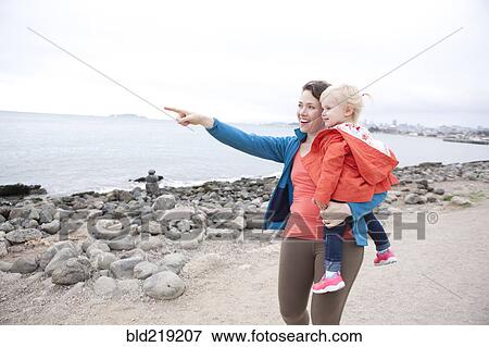 Stock Photo - Caucasian mother holding daughter and pointing on beach. Fotosearch