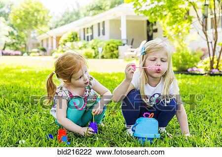 Stock Image - Caucasian sisters blowing bubbles in backyard. Fotosearch