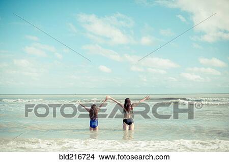 Picture - Caucasian sisters standing in waves on beach. Fotosearch