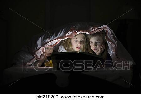 Stock Image - Caucasian sisters using laptop in blanket fort night. Fotosearch