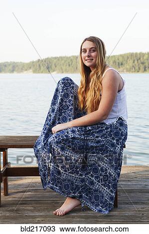 Stock Image - Caucasian teenage girl sitting on wooden dock at lake. Fotosearch