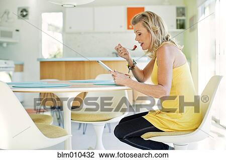 Stock Image - Caucasian woman eating and using digital tablet in dining room. Fotosearch