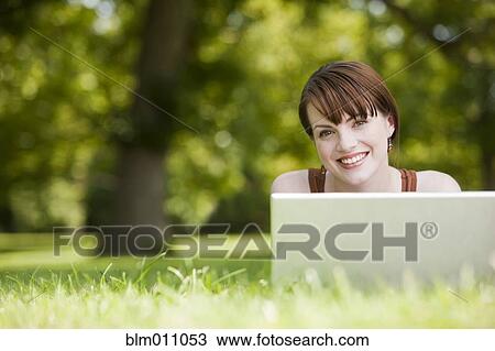 Stock Image - Caucasian woman laying in grass using laptop. Fotosearch
