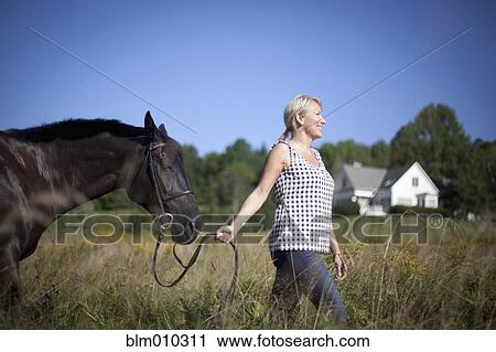 Stock Image - Caucasian woman leading horse through field. Fotosearch