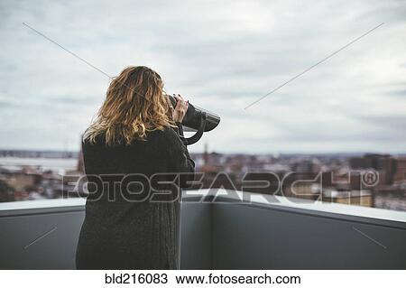 Stock Image - Caucasian woman on urban rooftop admiring cityscape with telescope. Fotosearch