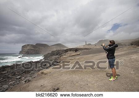 Stock Image - Caucasian woman photographing rocky beach. Fotosearch