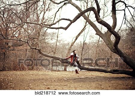 Stock Image - Caucasian woman sitting in tree in rural field. Fotosearch