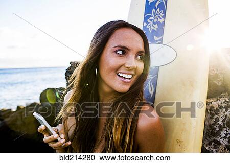 Stock Image - Caucasian woman using cell phone near surfboard. Fotosearch