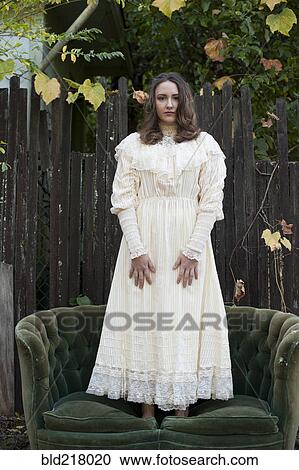 Stock Image - Caucasian woman wearing vintage gown standing on sofa. Fotosearch