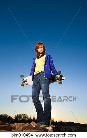 Picture - Chinese girl standing on rocks holding skateboard. Fotosearch
