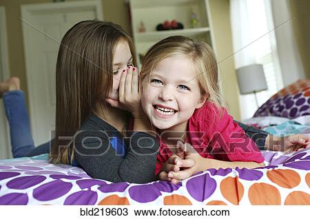 Stock Image - Close up of sisters whispering on bed. Fotosearch
