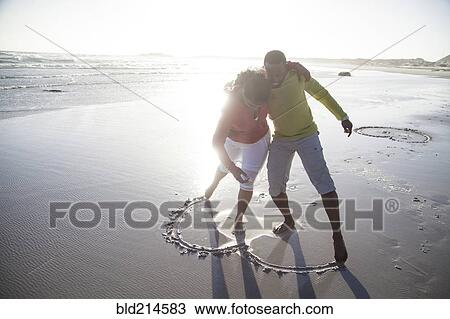 Stock Image - Couple drawing heart in sand on beach. Fotosearch
