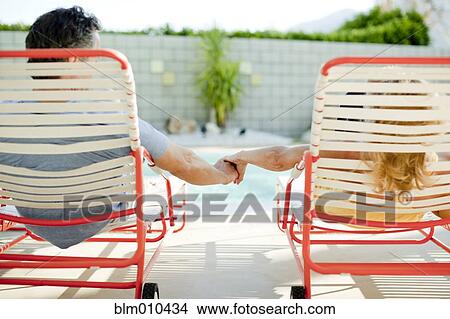 Picture - Couple holding hands in lounge chair near swimming pool. Fotosearch