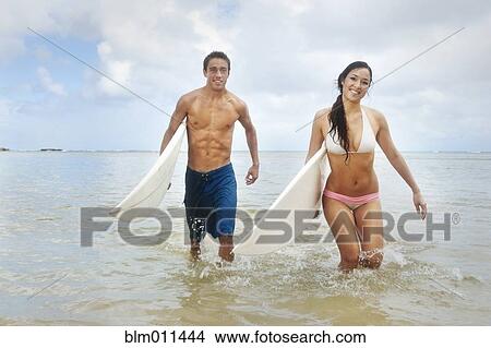 Picture - Couple walking in ocean carrying surfboards. Fotosearch