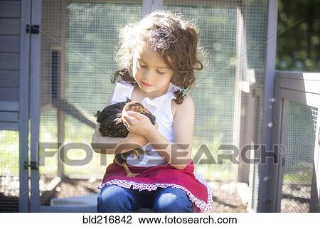 Stock Image - Curious girl holding chicken on farm. Fotosearch