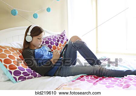 Stock Image - Girl listening to headphones with digital tablet on bed. Fotosearch
