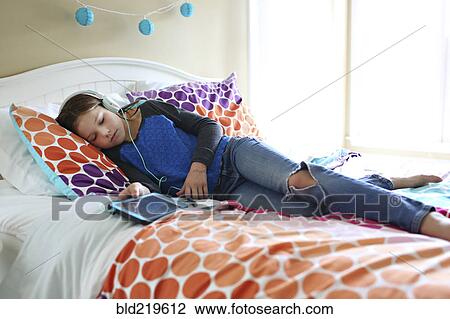 Stock Image - Girl listening to headphones with digital tablet on bed. Fotosearch