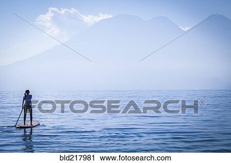 Stock Image - Girl standing on paddleboard in remote lake. Fotosearch