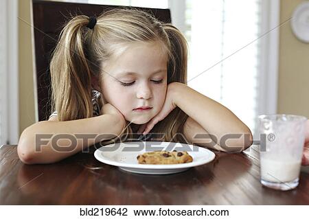 Stock Image - Girl staring at cookie with milk at table. Fotosearch