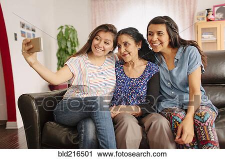 Stock Image - Grandmother and granddaughters taking selfie on sofa. Fotosearch