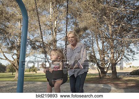 Stock Image - Grandmother pushing granddaughter on swing at playground. Fotosearch