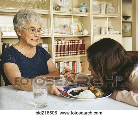 Stock Photograph - Grandmother serving granddaughter food at dining table. Fotosearch