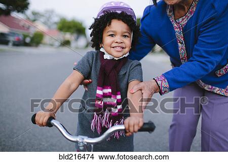 Picture - Grandmother teaching granddaughter to ride bicycle on suburban street. Fotosearch